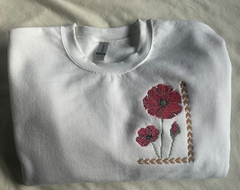May include: White sweatshirt with a red embroidered poppy flower design. The design includes a large poppy, a smaller poppy, and a bud. The design is surrounded by a brown embroidered border.