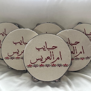 May include: A collection of six round tambourines with a beige fabric surface. Each tambourine features embroidered Arabic text in a deep red color, with a decorative floral border in red and black. The tambourines have silver jingles.