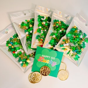 Könnte beinhalten: Fünf durchsichtige Plastiktüten gefüllt mit grünen, weißen und goldenen Perlen. Eine grüne Karte mit dem Text "Happy Go Lucky" in Regenbogenfarben. Vier goldene Münzen mit einem Kleeblatt-Design.