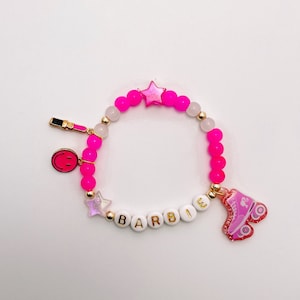 Puede incluir: Pulsera de cuentas rosa y blancas con dijes dorados, incluyendo una estrella, una carita sonriente, un labial y un pat&iacute;n. La pulsera tambi&eacute;n tiene cuentas blancas con la palabra "BARBIE" escrita.