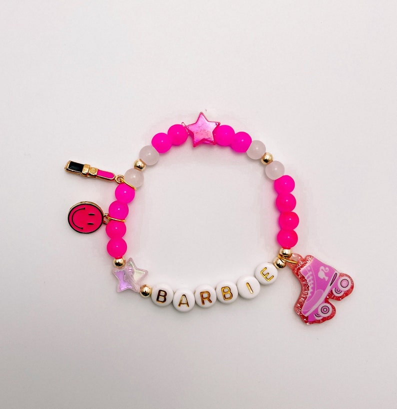 Puede incluir: Pulsera de cuentas rosa y blancas con dijes dorados, incluyendo una estrella, una carita sonriente, un labial y un pat&iacute;n. La pulsera tambi&eacute;n tiene cuentas blancas con la palabra "BARBIE" escrita.