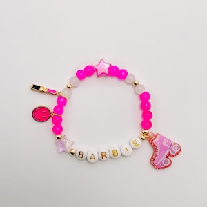 Puede incluir: Pulsera de cuentas rosa y blancas con dijes dorados, incluyendo una estrella, una carita sonriente, un labial y la palabra "BARBIE". La pulsera tambi&eacute;n tiene un dije de patines rosa.