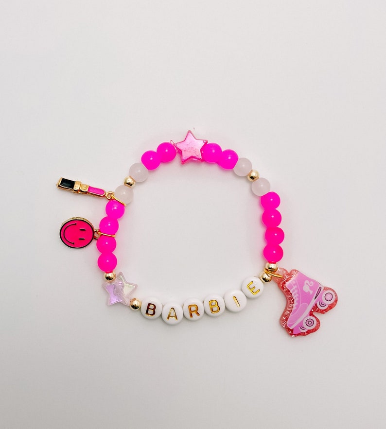 Puede incluir: Pulsera de cuentas rosa y blancas con dijes dorados, incluyendo una estrella, una carita sonriente, un labial y la palabra "BARBIE". La pulsera tambi&eacute;n tiene un dije de patines rosa.
