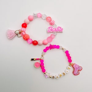 Puede incluir: Dos pulseras de cuentas rosas y blancas con dijes. La pulsera superior tiene un dije rosa de "Barbie" y una borla rosa. La pulsera inferior tiene un dije de patines rosa y un dije blanco de "Barbie".