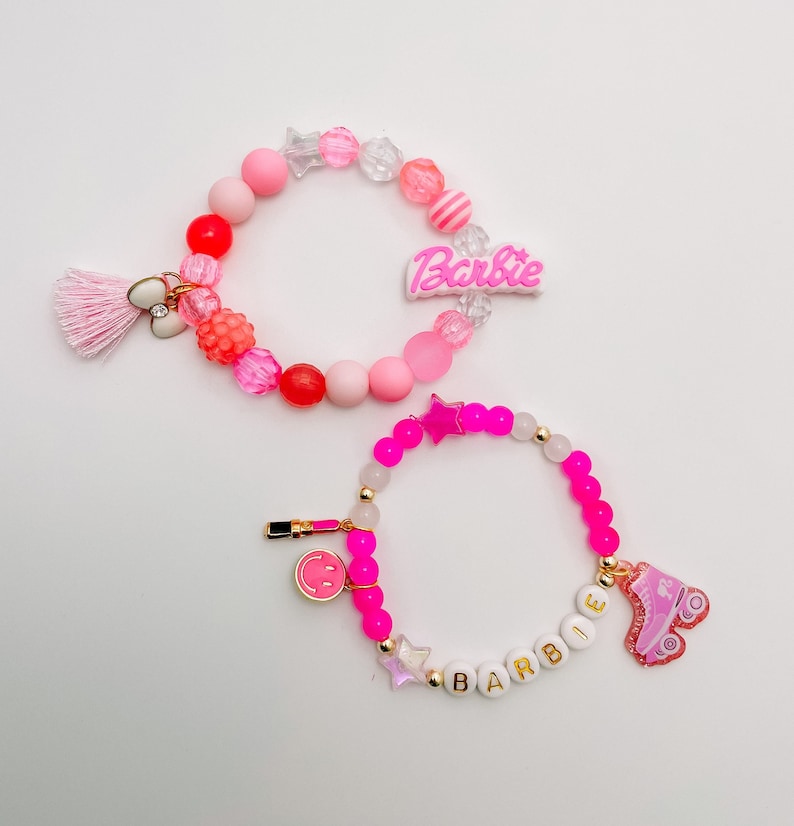 Puede incluir: Dos pulseras de cuentas rosas y blancas con dijes. La pulsera superior tiene un dije rosa de "Barbie" y una borla rosa. La pulsera inferior tiene un dije de patines rosa y un dije blanco de "Barbie".