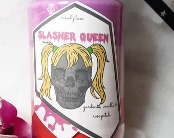 May include: A glass jar candle with a black lid, featuring a label with the text "SLASHER QUEEN" and a skull graphic. The candle has a layered appearance with pink and red hues. Decorative purple orchids and a black star-shaped object are also in the image.