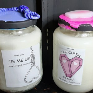 May include: Two glass jars filled with a white substance, likely candles. One jar has a blue and white twisted rope design on the lid, the other has a pink and black lid. Labels read "TIE ME UP" and "YOUR COFFIN, OR MINE?"