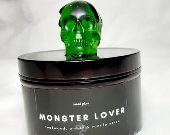 May include: A dark brown jar with a green skull-shaped lid. The jar has a black label with the words "MONSTER LOVER" and the scent description "teakwood, amber & vanilla spice". The text "inked plum" is also on the label.