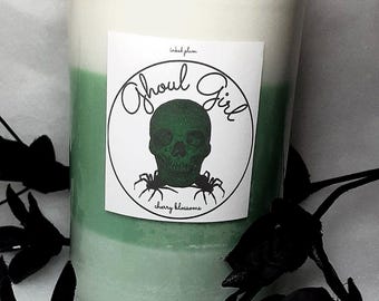 May include: A layered candle in a glass jar with a black lid. The candle has white, green, and light green layers. A label features a green skull and the words "Ghoul Girl" and "cherry blossoms". Black decorative elements surround the candle.