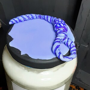 May include: A glass jar candle with a light purple lid and a blue rope design. The candle label reads "TIE ME UP" with a noose illustration. The candle is scented with brown sugar and cinnamon.