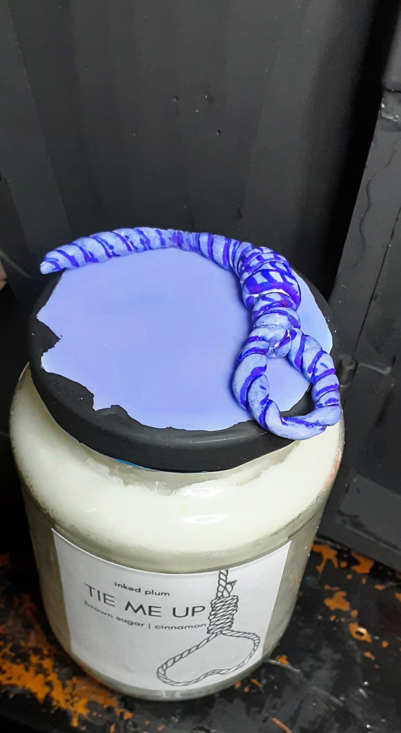May include: A glass jar candle with a light purple lid and a blue rope design. The candle label reads "TIE ME UP" with a noose illustration. The candle is scented with brown sugar and cinnamon.