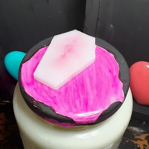 May include: A white candle jar with a black lid, topped with a pink coffin-shaped wax piece. The lid has a vibrant pink paint design. The label reads "YOUR COFFIN OR MINE?"