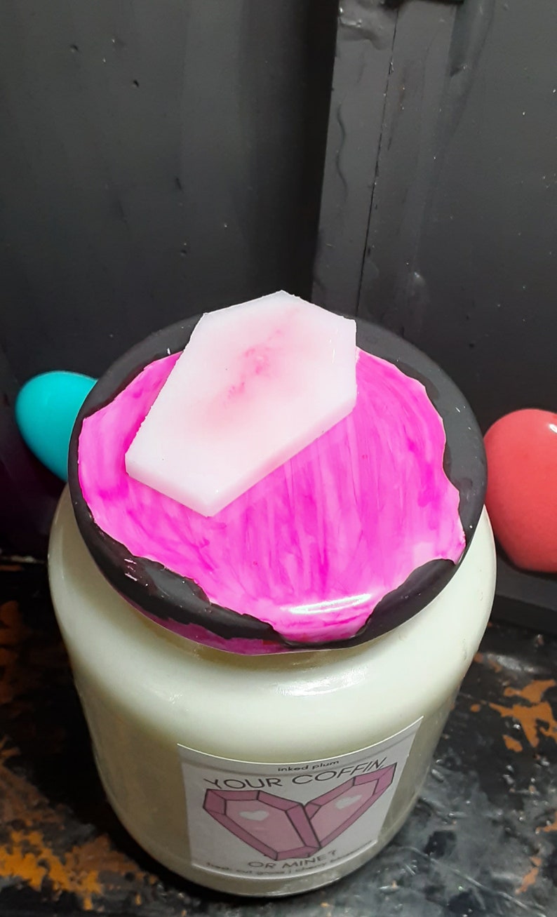 May include: A white candle jar with a black lid, topped with a pink coffin-shaped wax piece. The lid has a vibrant pink paint design. The label reads "YOUR COFFIN OR MINE?"