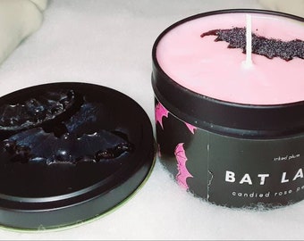 May include: A black tin candle with a pink wax interior and a bat-shaped design. The candle is labeled "BAT LADY" with "candied rose petals" below. The lid is off to the side, revealing a bat design.