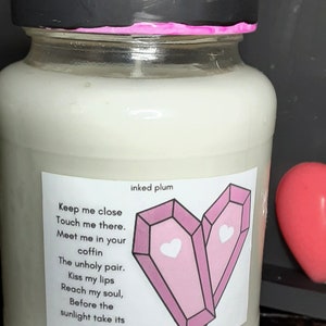 May include: A glass jar candle with a black lid and a pink rim. The label features a heart-shaped design of two pink coffins with hearts, and the text "inked plum" and a poem. A small, light-colored lid sits on top.