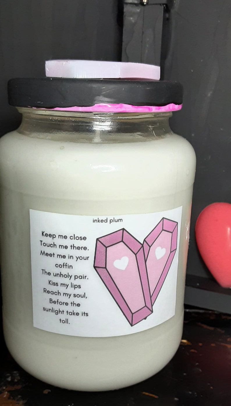 May include: A glass jar candle with a black lid and a pink rim. The label features a heart-shaped design of two pink coffins with hearts, and the text "inked plum" and a poem. A small, light-colored lid sits on top.