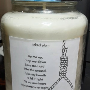 May include: A glass jar candle with a black lid and a blue decorative element on top. The label reads "inked plum" and features a poem and a noose illustration. The candle is filled with a creamy wax.