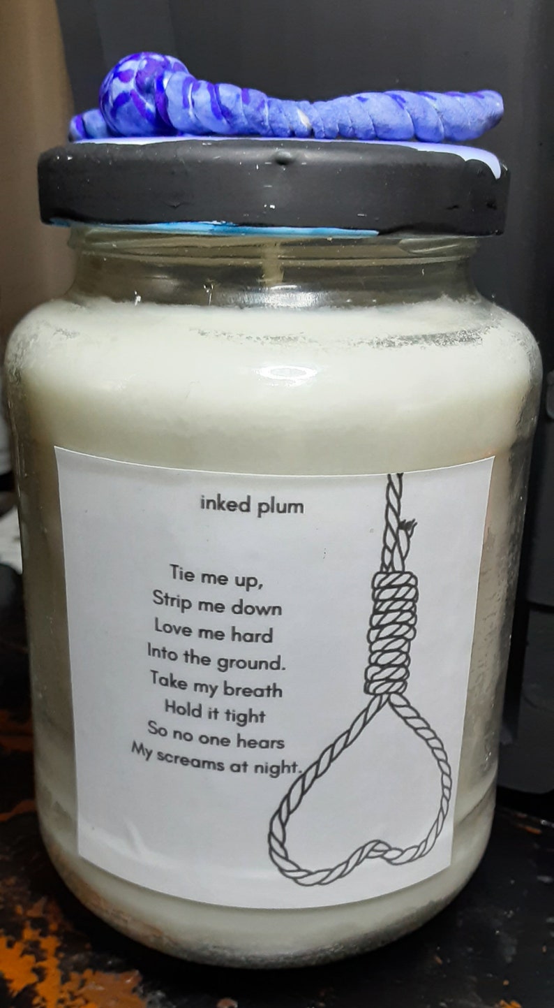 May include: A glass jar candle with a black lid and a blue decorative element on top. The label reads "inked plum" and features a poem and a noose illustration. The candle is filled with a creamy wax.