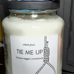 May include: A glass jar candle with a black lid and a decorative blue and purple swirl on top. The label reads "TIE ME UP" with a noose graphic, and lists "brown sugar | cinnamon" as scents.