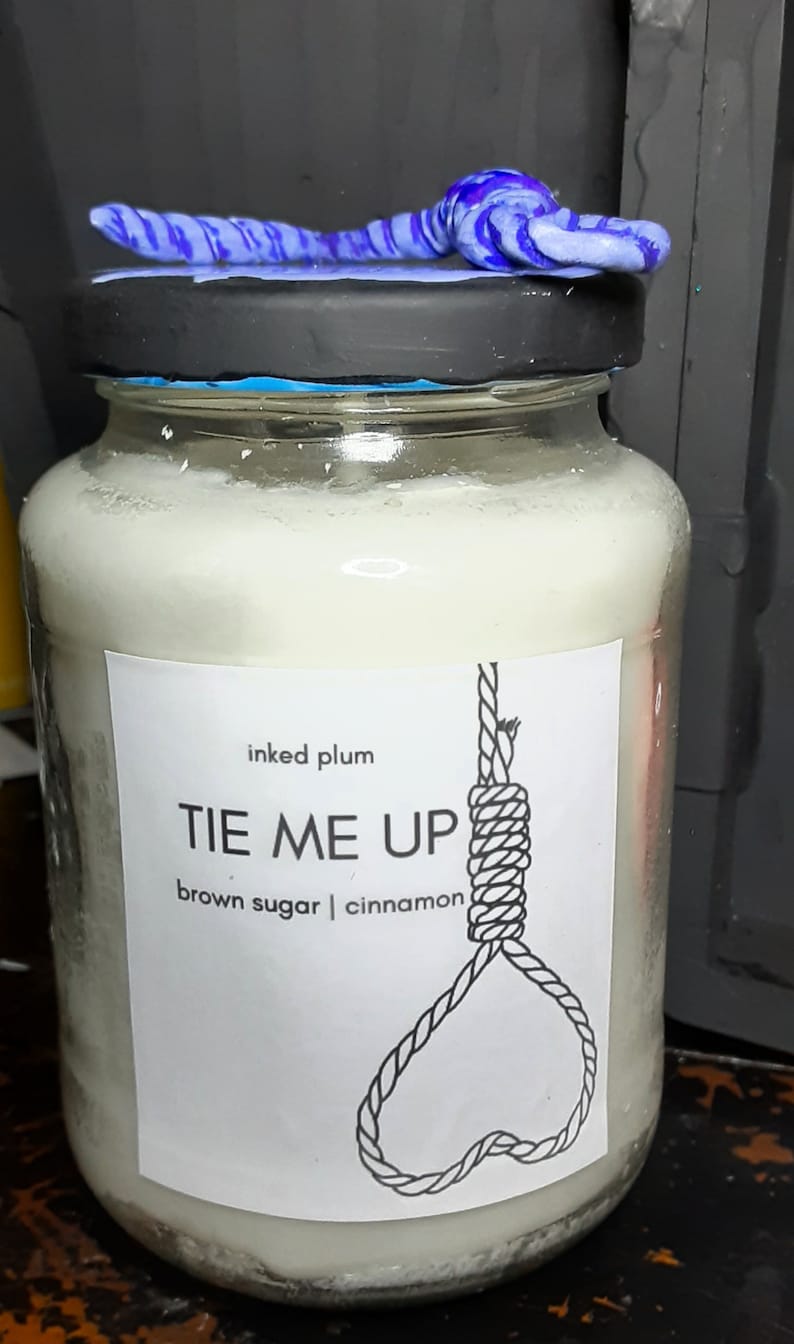 May include: A glass jar candle with a black lid and a decorative blue and purple swirl on top. The label reads "TIE ME UP" with a noose graphic, and lists "brown sugar | cinnamon" as scents.