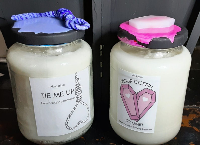 May include: Two glass jars filled with a white substance, likely candles. One jar has a blue and white twisted rope design on the lid, the other has a pink and black lid. Labels read "TIE ME UP" and "YOUR COFFIN, OR MINE?"