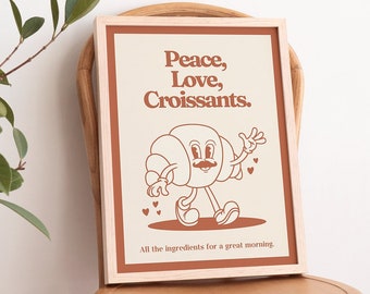 May include: A framed print with a light brown background and a brown border. The print features a cartoon croissant with a mustache and a smile, walking and holding a heart. The text on the print reads "Peace, Love, Croissants. All the ingredients for a great morning."