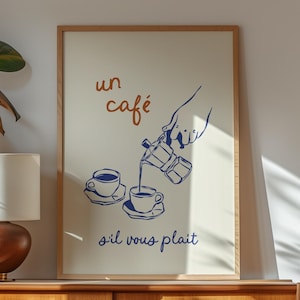 以下が含まれることがあります： モカポットから2つのカップにコーヒーを注ぐ手のミニマルなイラストが描かれた額装されたプリント。"un café s'il vous plaît"というテキストが青とオレンジで書かれています。