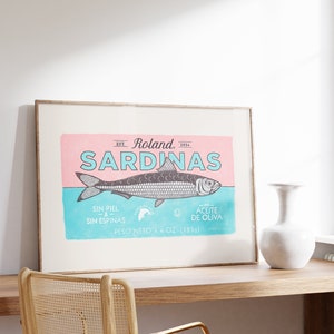 May include: A framed vintage-style poster with a pink and blue background featuring a sardine illustration and the text "EST. Roland, 1934 SARDINAS SIN PIEL -&- SIN ESPINAS 0 -en- ACEITE DE OLIVA PESO NETO 4.4 OZ. (125g)"