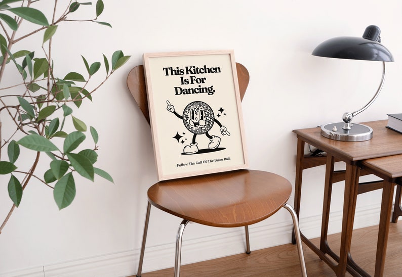 May include: A framed print with the text "This Kitchen Is For Dancing" and a cartoon disco ball character. The print is displayed on a wooden chair. The image has a retro, vintage aesthetic.