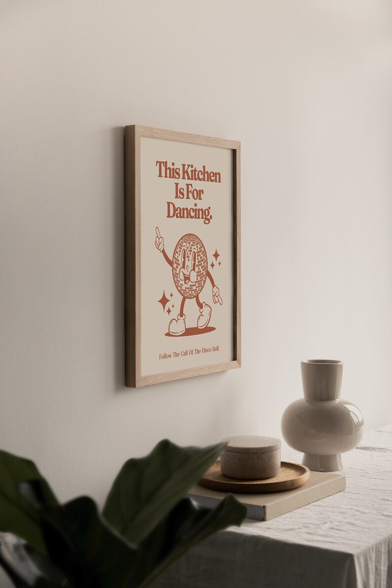 May include: A framed print with a brown background and a cartoon disco ball character dancing. The text reads "This Kitchen Is For Dancing. Follow The Call Of The Disco Ball."