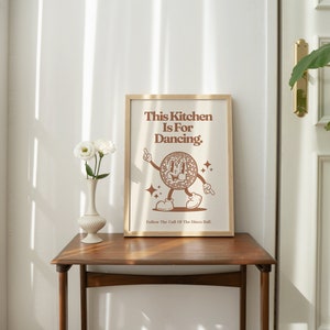 May include: A brown framed print with the text "This Kitchen Is For Dancing." and a cartoon illustration of a dancing cookie. The print is hanging on a white wall above a wooden table.