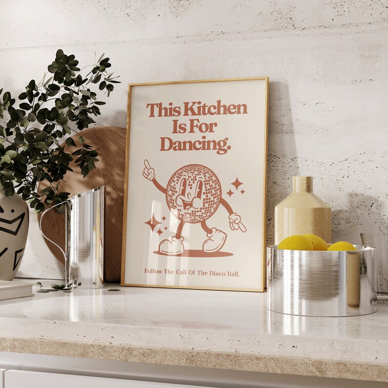 May include: A gold framed poster with a white background and red text that reads "This Kitchen Is For Dancing." A cartoon disco ball with a smiling face is dancing. The text at the bottom reads "Follow The Call Of The Disco Ball."