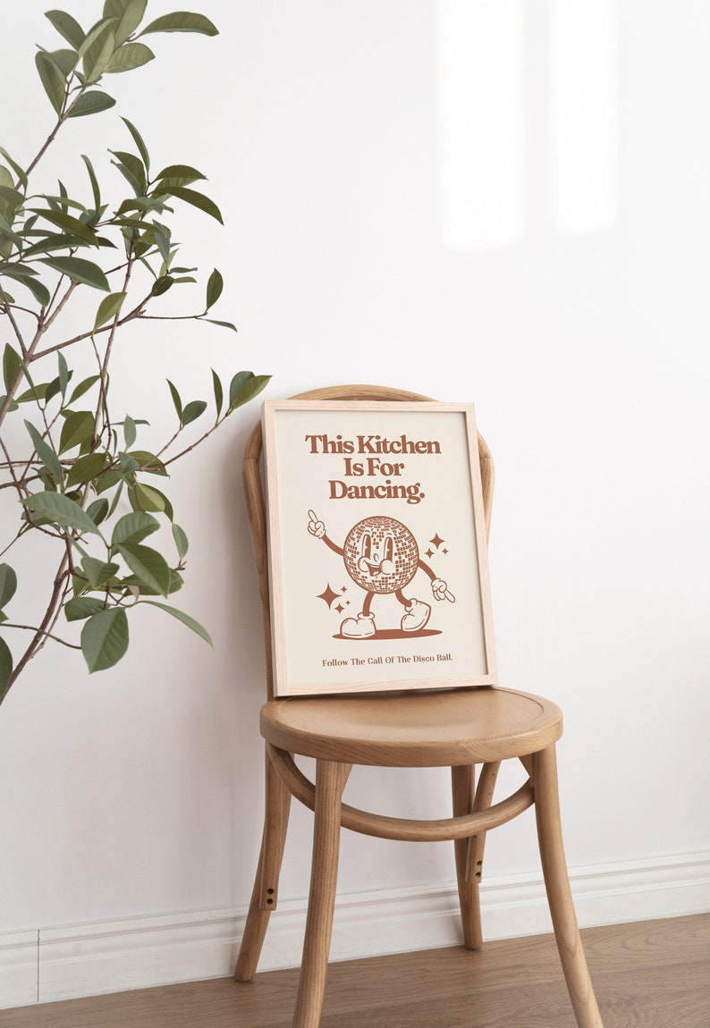 May include: A framed print with a brown background and white text that reads "This Kitchen Is For Dancing." A cartoon disco ball with a smiling face is dancing with its arms outstretched. The text "Follow The Call Of The Disco Ball." is at the bottom of the print.
