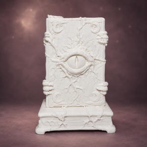 May include: A white rectangular book-shaped sculpture featuring a central sculpted eye, vine-like textures, and clawed hands gripping the sides. The cover displays crescent moon motifs in the corners, and the entire piece rests on a base.