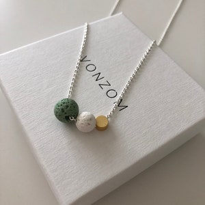 Könnte beinhalten: Eine silberne Kettenkette mit drei Perlen: eine grüne Lavaperle, eine weiße Lavaperle und eine goldene Perle. Die Kette liegt auf einer weißen Schachtel mit dem aufgedruckten Wort "VONZO".