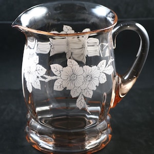以下が含まれることがあります： 花柄のピンク色のガラス製のピッチャー。ピッチャーは口が広く、持ち手があります。花柄はガラスにエッチングされており、白い花と葉が描かれています。