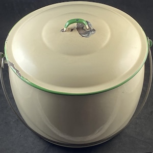 May include: A cream-colored enamel bucket with a matching lid, featuring a thin green rim and a green handle on the lid. The bucket has a metal swing handle attached to the sides, with visible signs of wear and chipping along the edges.