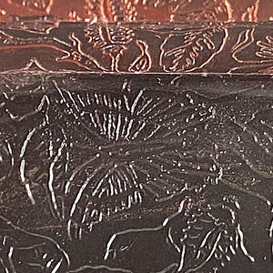 May include: Close-up of a dark brown, textured surface with embossed floral and animal designs. The image shows a detailed pattern with raised relief, creating a tactile and visual contrast. The design includes leaves and animal figures.