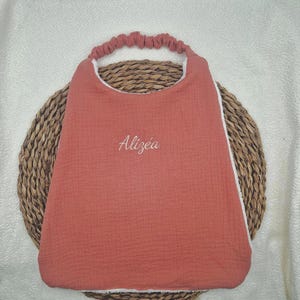 Peut inclure: Un bavoir pour bébé de couleur terre cuite avec un tour de cou élastique froncé et le prénom Alizéa brodé en blanc. Le bavoir présente une face en tissu texturé et un envers blanc, offrant un design simple et fonctionnel au quotidien.