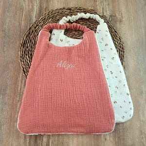 Peut inclure: Deux bavoirs pour bébé avec encolure élastique. Le bavoir avant est en mousseline unie de couleur terre cuite avec le prénom Alizéa brodé en fil blanc. Le bavoir arrière est en mousseline blanc cassé avec un motif floral de petites fleurs.