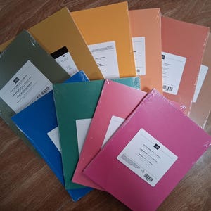 May include: A collection of rectangular paper packs arranged in a fan shape. Each pack is wrapped in clear plastic and features a white rectangular label. The paper colors include olive green, mustard yellow, orange, coral, pink, teal, and blue.