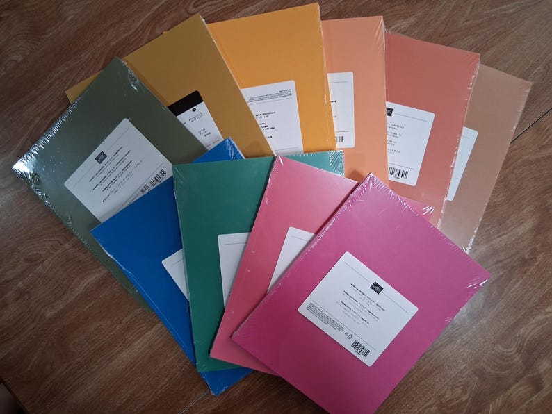 May include: A collection of rectangular paper packs arranged in a fan shape. Each pack is wrapped in clear plastic and features a white rectangular label. The paper colors include olive green, mustard yellow, orange, coral, pink, teal, and blue.