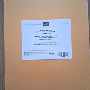 May include: A pack of 24 sheets of Mango Melody cardstock measuring 8-1/2 inches by 11 inches, sealed in clear plastic. The white label features the Stampin' Up! logo, product name, quantity, and dimensions in both English and French.