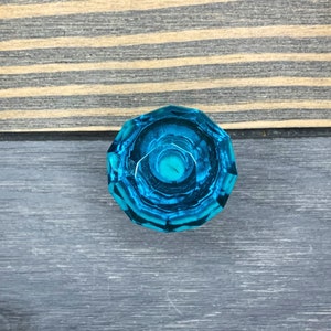 May include: A faceted, teal-colored glass knob. The knob is round with a detailed, geometric design. It is set against a backdrop of wooden planks in shades of brown and gray.