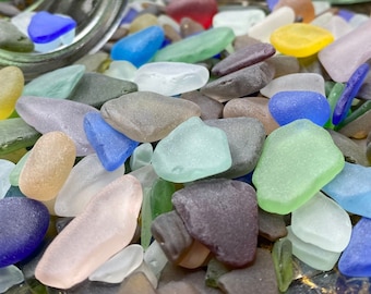 May include: A close-up of a glass jar filled with colorful sea glass. The jar is tipped over, spilling the sea glass onto a wooden surface. The sea glass is a variety of colors, including blue, green, brown, and white.