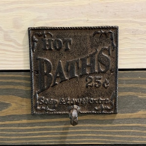 May include: A cast iron sign with the text "Hot Baths 25¢ Soap & Towels Extra" on a wooden wall.