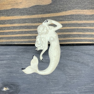 May include: A white ceramic mermaid figurine with black eyes and a black tail fin. The mermaid is posed with her hand on her head and her tail curled behind her.