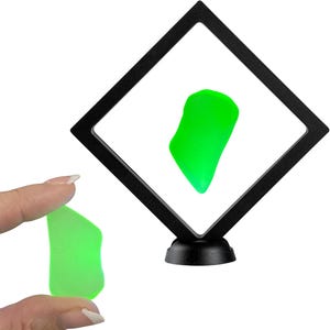 May include: A vibrant green sea glass piece, held by a hand, next to a black display case with a similar piece inside. The sea glass has a smooth, irregular shape and a bright, translucent color.