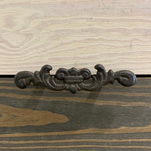 May include: A black metal drawer pull with a decorative design. The pull is mounted on a wooden surface with a rustic finish.