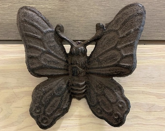 May include: A cast iron butterfly wall hook in a dark brown color. The hook is shaped like a butterfly with its wings spread out. It is a decorative and functional piece for hanging items on a wall.